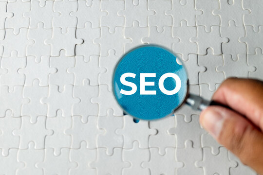 Hand holding a magnifying glass over a white jigsaw puzzle revealing the word SEO. Concept of Search Engine Optimization, digital marketing strategy, and internet business solutions.