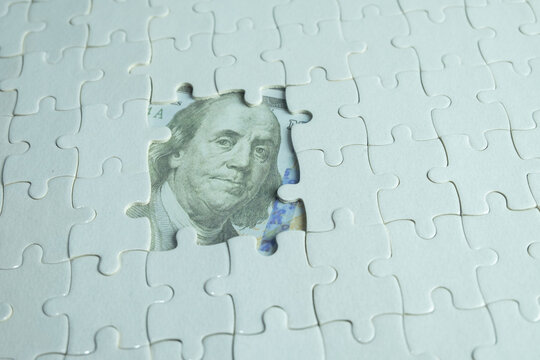 White jigsaw puzzle with a missing section revealing a 100 US dollar bill underneath. Concept of financial solution, business strategy, hidden wealth, and solving economic problems.