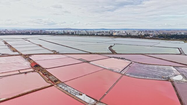 Salt extraction is underway in a grid of pink and red evaporation ponds, with dikes dividing the water and city buildings visible on the horizon.