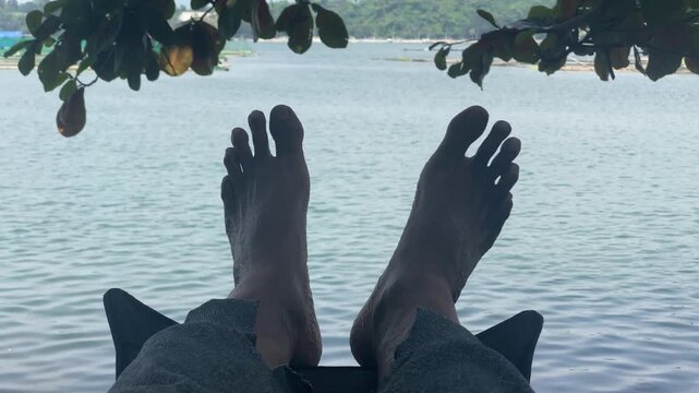 old man sits in a low-profile camping chair with his bare feet dangling above a calm body of water, framed by tree branches overhead.