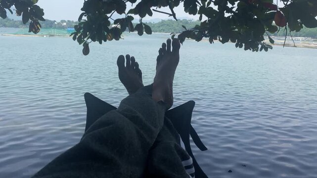 old man sits in a low-profile camping chair with his bare feet dangling above a calm body of water, framed by tree branches overhead.