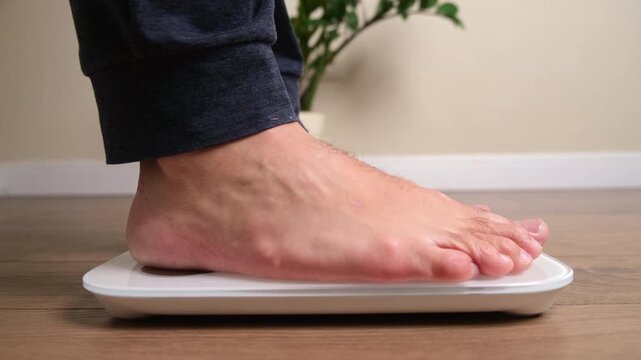 Close up of bare foot of man standing on white digital bathroom scale on wooden floor. Weight control and body mass measurement concept for healthy lifestyle and diet monitoring
