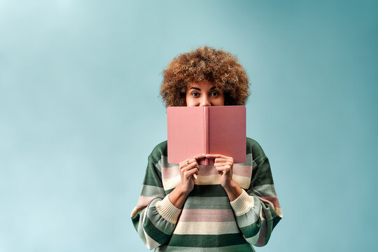 Young person with curly hair holds a pink book over their lower face, leaving only the eyes visible, wearing a striped sweater against a soft turquoise background.