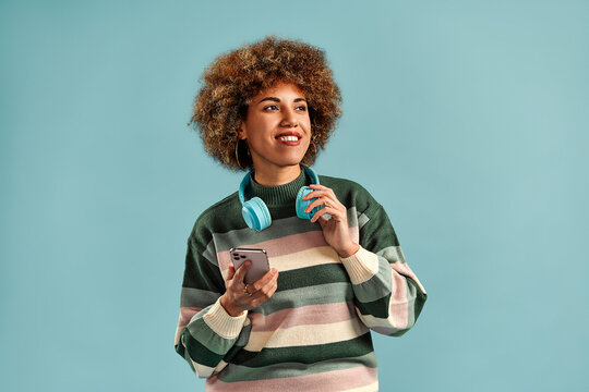 Smiling young woman with curly hair holds a smartphone and wears teal headphones against a plain blue studio background with copy space.