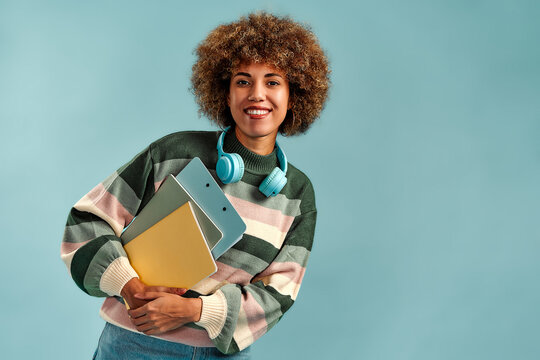 Smiling young woman with curly hair holding notebooks and folders, wearing headphones and a striped sweater against a blue studio background.
