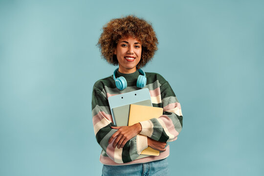 Young woman with curly hair, headphones around her neck, and books in her arms, standing against a blue background in a bright student portrait.