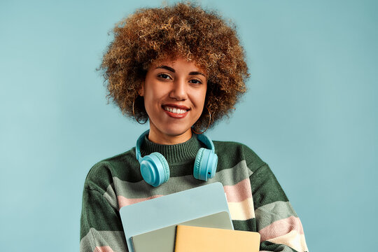 Portrait of young woman with curly hair wearing headphones around her neck and holding colorful folders against a plain turquoise background.