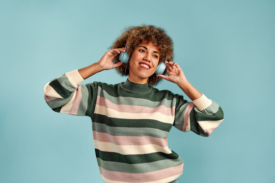 Beautiful young woman with curly hair wearing headphones and a striped sweater, enjoying music against a plain blue studio background.