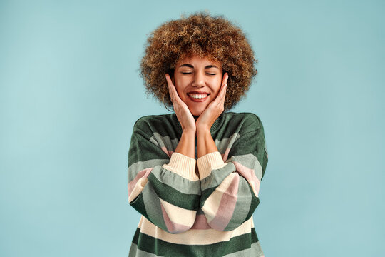 Smiling young woman with curly hair and closed eyes touching her cheeks, wearing a striped sweater against a light blue studio background.