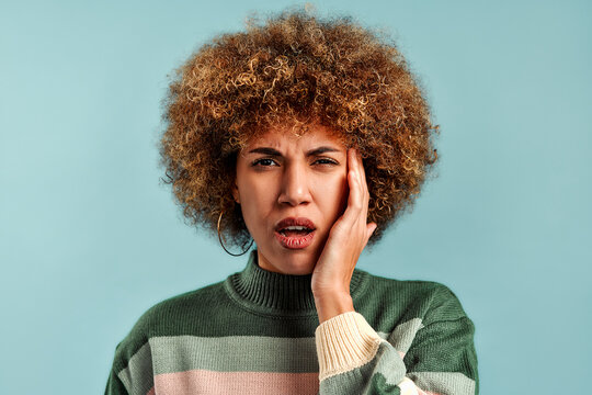 Portrait of a young woman with curly hair holding her cheek and looking worried against a light blue studio background.