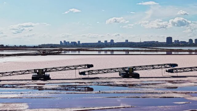 Salt extraction takes place in shallow ponds, where conveyor equipment crosses the water and workers tend the site near a city skyline during the day.