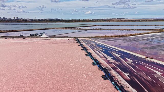 Salt extraction ponds cover a coastal site, with pink water, salt beds, and channels visible during daylight beneath a cloudy sky.