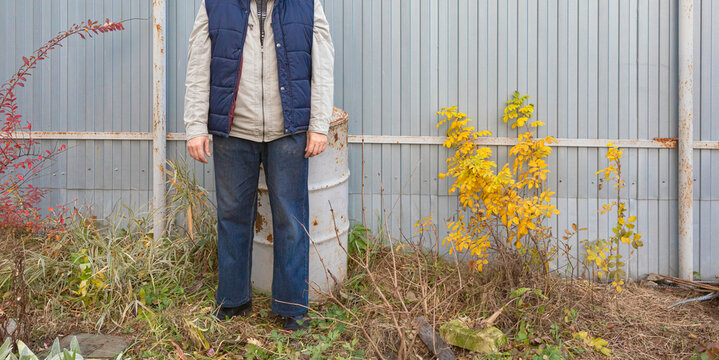 Cropped view of person standing among overgrown plants beside a weathered metal fence. Autumn foliage and industrial garden surroundings. Outdoor portrait with copy space