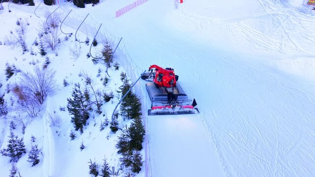 Snowcat grooming ski slope near pine trees, Pamporovo, Bulgaria. Red snow groomer tractor leveling fresh snow track beside safety net and forest.