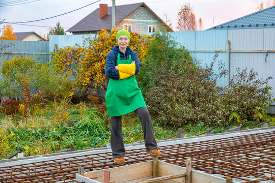Woman in gardening apron stands on rebar at home construction site. Residential building work and garden renovation. DIY home improvement and outdoor project. Construction and gardening