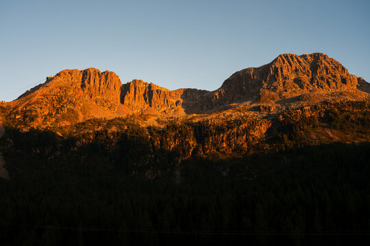 Majestic mountain peaks glowing in warm golden sunset light above dark pine forest in Dolomites, Italy.