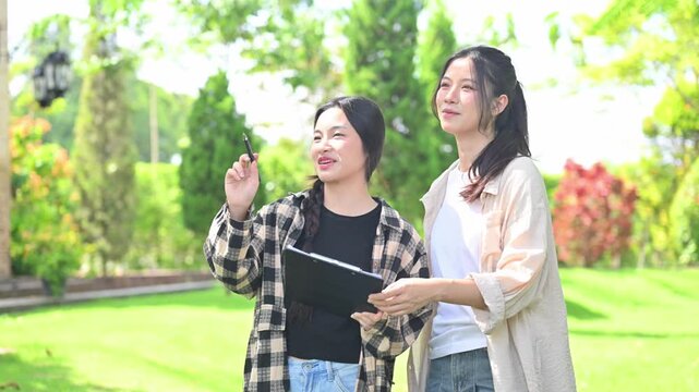 Two Young Women Reviewing Clipboard Documents Together in a Sunny Green Park for Agriculture Field Research