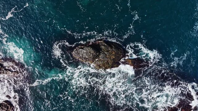 White surf washes over dark rocks as deep blue and turquoise water moves around a small outcrop near the shore in daylight.