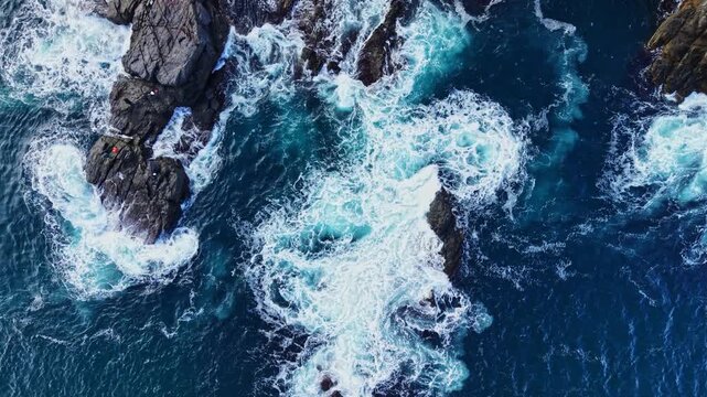 From above, blue ocean waves surge and foam around dark rocks along a rugged coast, with white water filling the gaps between the formations.