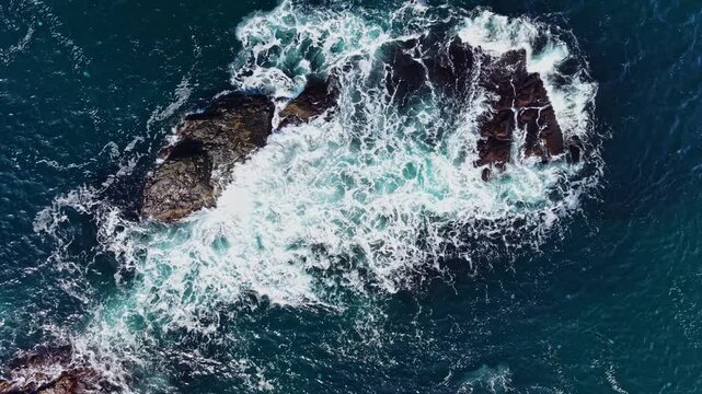 White surf moves around exposed rocks as dark blue seawater flows and breaks across the rocky coastline in daylight.