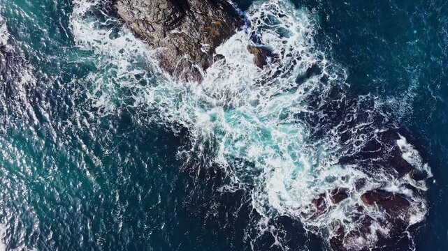 Water churns and forms white foam as waves move across rocks along the coast, seen from above in daylight.