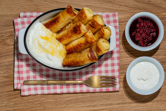 Stuffed crepes pancakes with sour cream and berry jam. Golden thin pancakes rolls in ceramic dish. Traditional blini or filled crepes breakfast served with sour cream, honey and raspberry jam