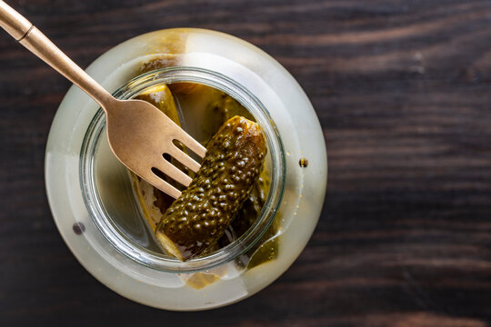 Pickled cucumber on golden fork from glass jar top view. Crunchy marinated gherkin on brass fork inside open jar. Traditional preserved organic cucumbers appetizer on wooden table for culinary menu