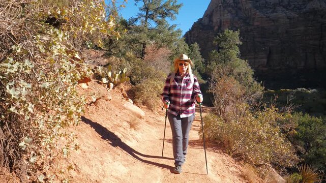 Senior woman wearing sun hat walking along dirt hiking trail with trekking poles under bright sunlight in zion national park. Hiker enjoying active retirement outdoor lifestyle on canyon path