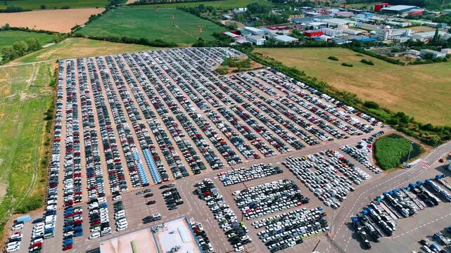 Fields surround a paved vehicle yard divided by car storage rows. Parked vehicles form long lines around access lanes and a service building.