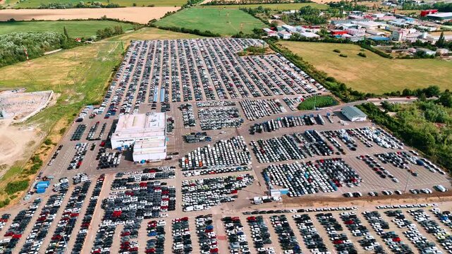 Industrial buildings and fields border rows of stored vehicles. Parallel vehicle rows surround a service building, with greenery along the site boundary.