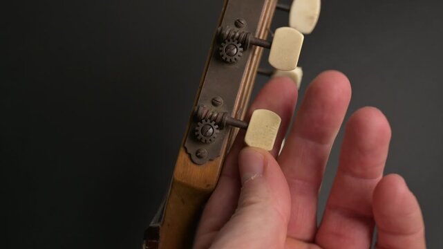 Hand adjusts an exposed geared tuning machine on the wooden headstock of a stringed instrument