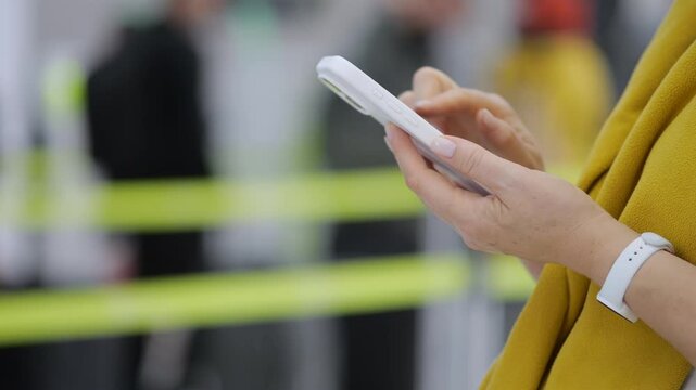 Person uses a smartphone while standing near yellow barriers. Their hands hold the phone with care. Background shows blurred people and barriers. The person wears a yellow jacket and white wristband