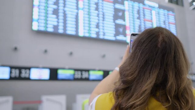 Woman with long brown hair wears yellow top. She holds phone up to large airport departure screen. Screen shows flight details and status updates. Background includes smaller digital display panels