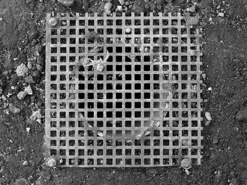 A top-down, high-contrast black and white photograph of a square metal grating cover bolted over a drain opening. The grid pattern of the drainage grate is set against rough asphalt or dark soil scatt