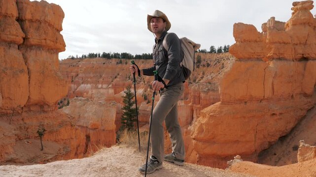 Man hiker with backpack standing on rocky cliff edge holding trekking poles and looking around sandstone formations in bryce canyon national park. Hiker standing to resting and calm breathing