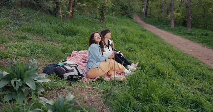 Two young women sitting on gingham blanket in woodland clearing reading and pointing toward treetop canopy while backpacks rest beside them on grass, relaxed overcast spring day in parkland scene