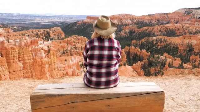 Female tourist wearing cowboy hat sitting on wooden bench, contemplating vast landscape of bryce canyon national park during sunny summer vacation trip. A fairy tale landscape created by nature.