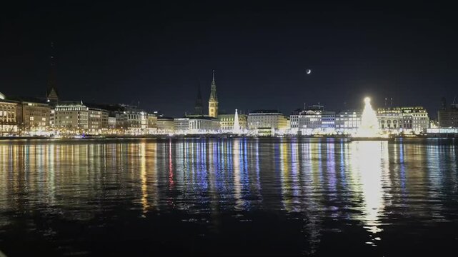 Timelapse of festive Alstertanne christmas tree illuminating Hamburg Binnenalster lake at night with crescent moon moving across sky