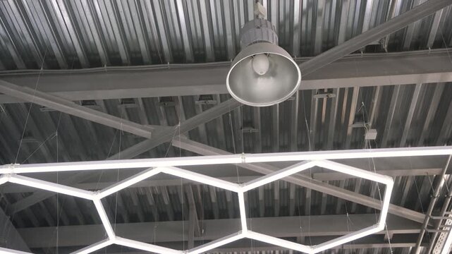 Camera tilts up focusing on a contemporary honeycomb shaped led lamp. The fixture hangs in a modern office or commercial space with an industrial style metal ceiling and steel beams