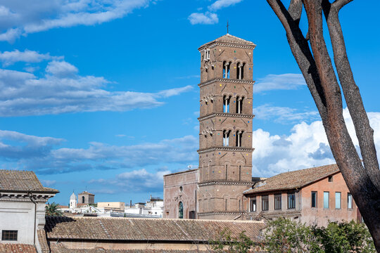 Medieval brick bell tower with Romanesque arched windows above terracotta rooftops and pine tree in Rome, Italy