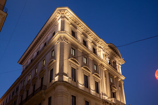 Illuminated neoclassical corner building at dusk with statues and ornate cornice in Rome, Italy