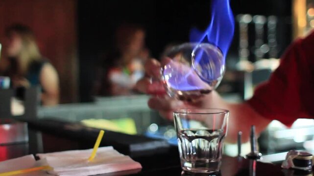 Bartender warming snifter glass with flaming sambuca cocktail over shot glass at nightclub bar