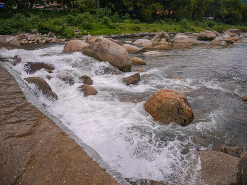 Rushing white water rapids and foam flowing vigorously over a concrete weir structure and smooth river boulders, depicting strong water current and river management.