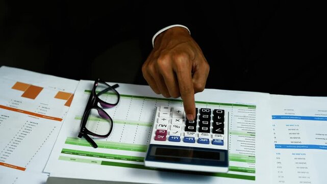 Person Calculating Budget on Financial Documents, Close-up of a person using a calculator on financial documents with charts, tables and eyeglasses, showing budgeting, accounting and tax planning.