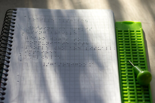 Horizontal stock photo of green braille slate and stylus next to open embossed Braille spiral notebook