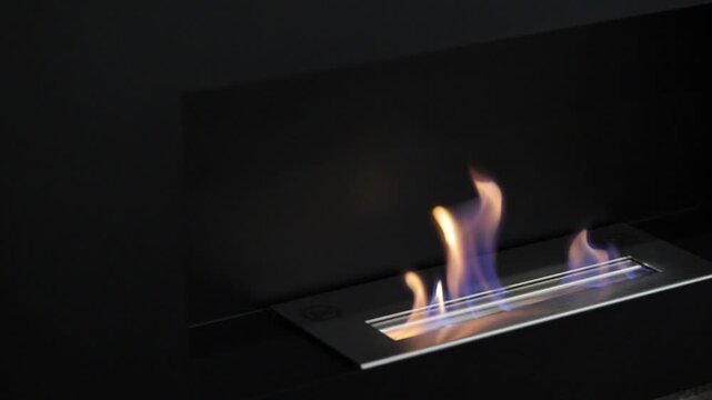 Diagonal camera movement showing sleek steel linear bio fireplace with fire tongues burning against dark minimal background inside contemporary apartment