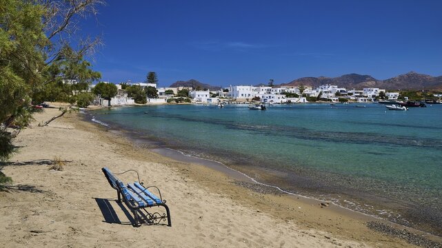 Empty bench on sandy beach overlooking the blue sea and a white coastal village, volcanic island, island, Milos, Cyclades, Greek Islands, Greece