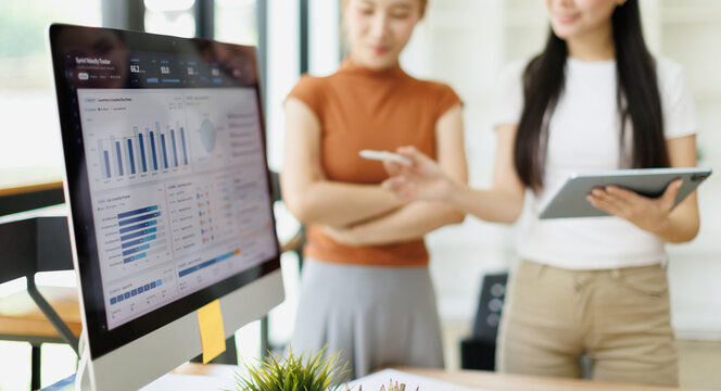 Business analysts discussing performance metrics, one pointing to charts on a desktop dashboard and a colleague holding a tablet during collaborative office planning