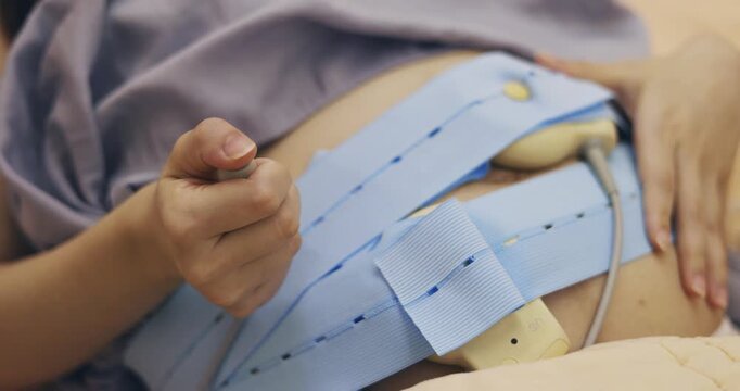 A pregnant woman lies in a hospital bed, using an instrument to detect the fetal heartbeat.