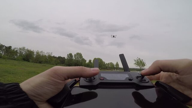POV close up pilot hands using joystick on remote controller to fly quadcopter drone over green field under cloudy sky